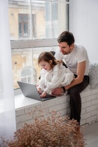 Father and daughter are playing on laptop, sitting on wooden, white windowsill near the window